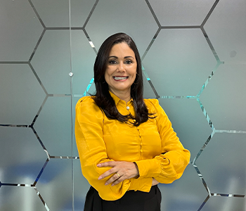 Coordenadora do curso de Direito, Soane Lopes, está de braços cruzados em frente ao um painel com desenhos em colmeia, camisa amarela, calça preta. Ela olha fixamente para a câmera e sorri.