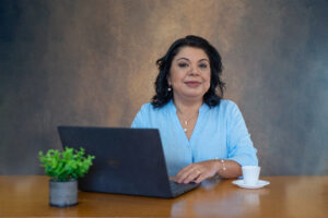 Camille Miguel, Diretora de Tecnologia e Inovação Digital da Clariens Educação sentada atrás de uma mesa de madeira mexendo no computador. Olhando para câmera e sorrindo, tem cabelos pretos e curtos, usa camisa azul formal, colar, brincos e pulseira. Tem também na foto uma xicara branca de café e um pequeno vazo com um planta.